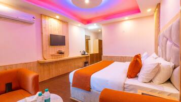 Una habitación de hotel con una cama, dos sillones naranjas, una mesita con botellas, un televisor montado en la pared y un mostrador de recepción de madera.