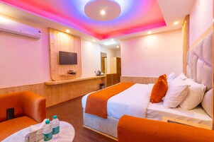 Una habitación de hotel con una cama, dos sillones naranjas, una mesita con botellas, un televisor montado en la pared y un mostrador de recepción de madera.