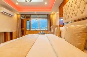 A hotel room with a large bed, a wooden headboard, a small table with a bottle and glasses, and a view of mountains through the window.