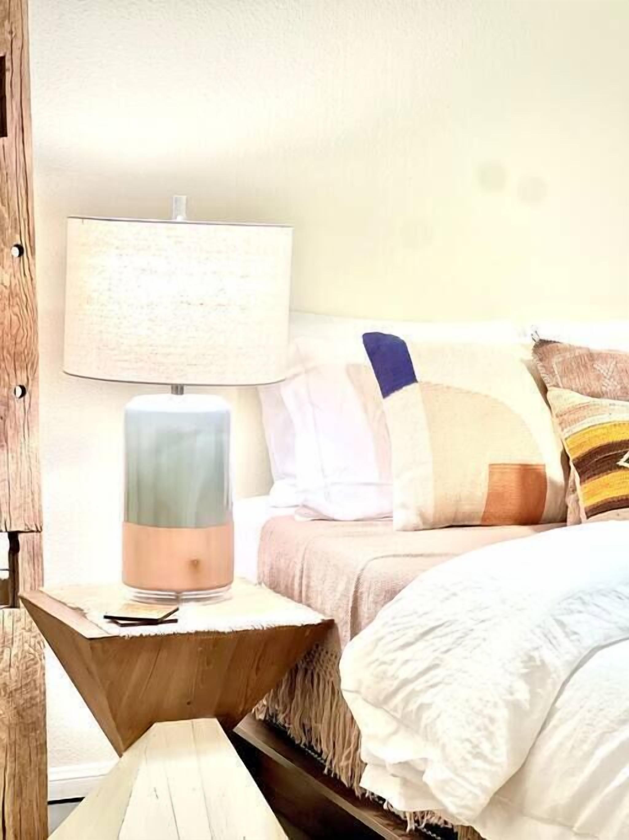 A bed with a white comforter and multiple pillows, a bedside table with a lamp, and a wooden nightstand.