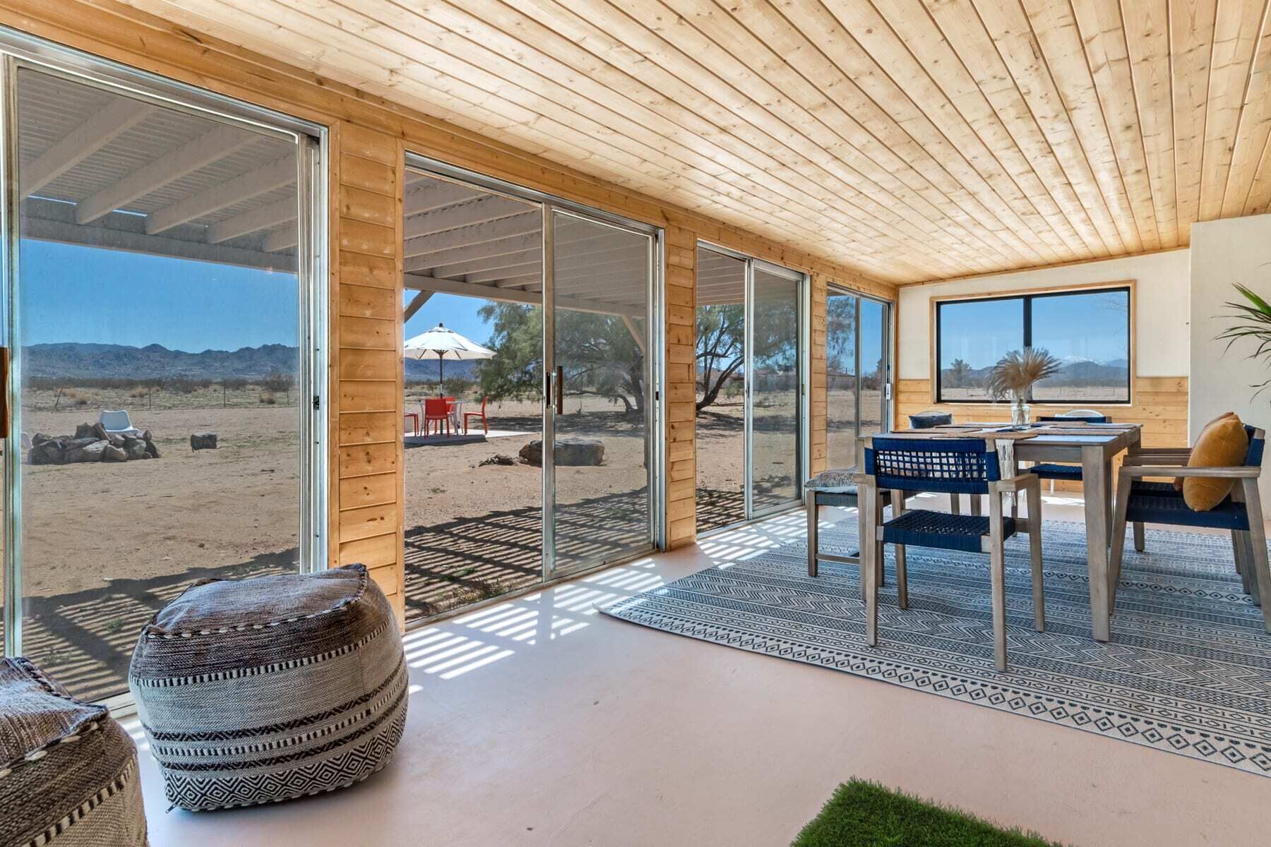 A modern interior with a large sliding glass door leading to a desert view, a wooden ceiling, and a dining area with a table and chairs.