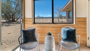 A room with two metal chairs, a small white table with a plant, and a large window offering a view of a desert landscape. - The Castle House: Hideaway w Cowboy Pool (Joshua Tree)