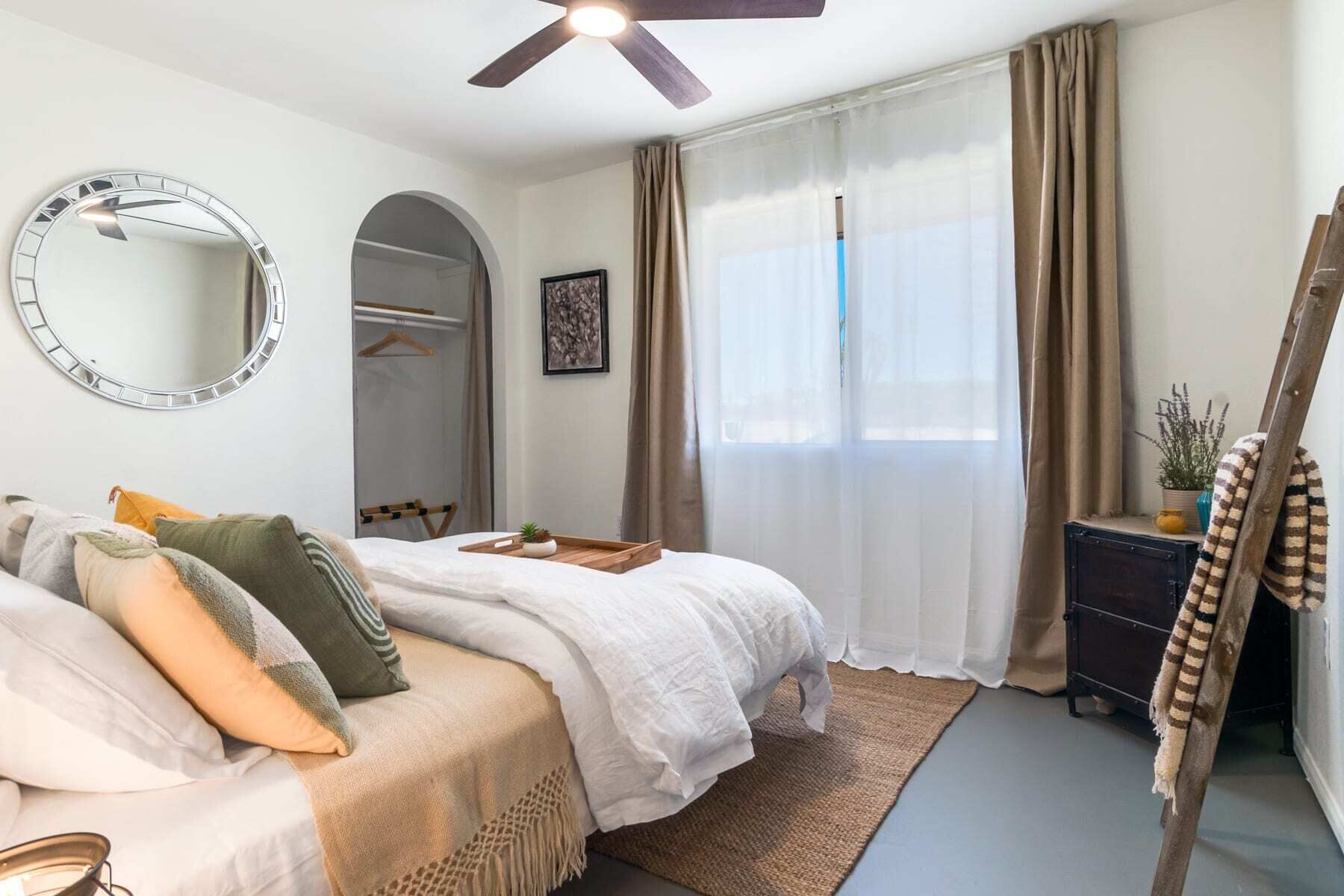 Un dormitorio con cama, un espejo redondo, ventilador de techo, ventana con cortinas, armario y una mesita con una planta.