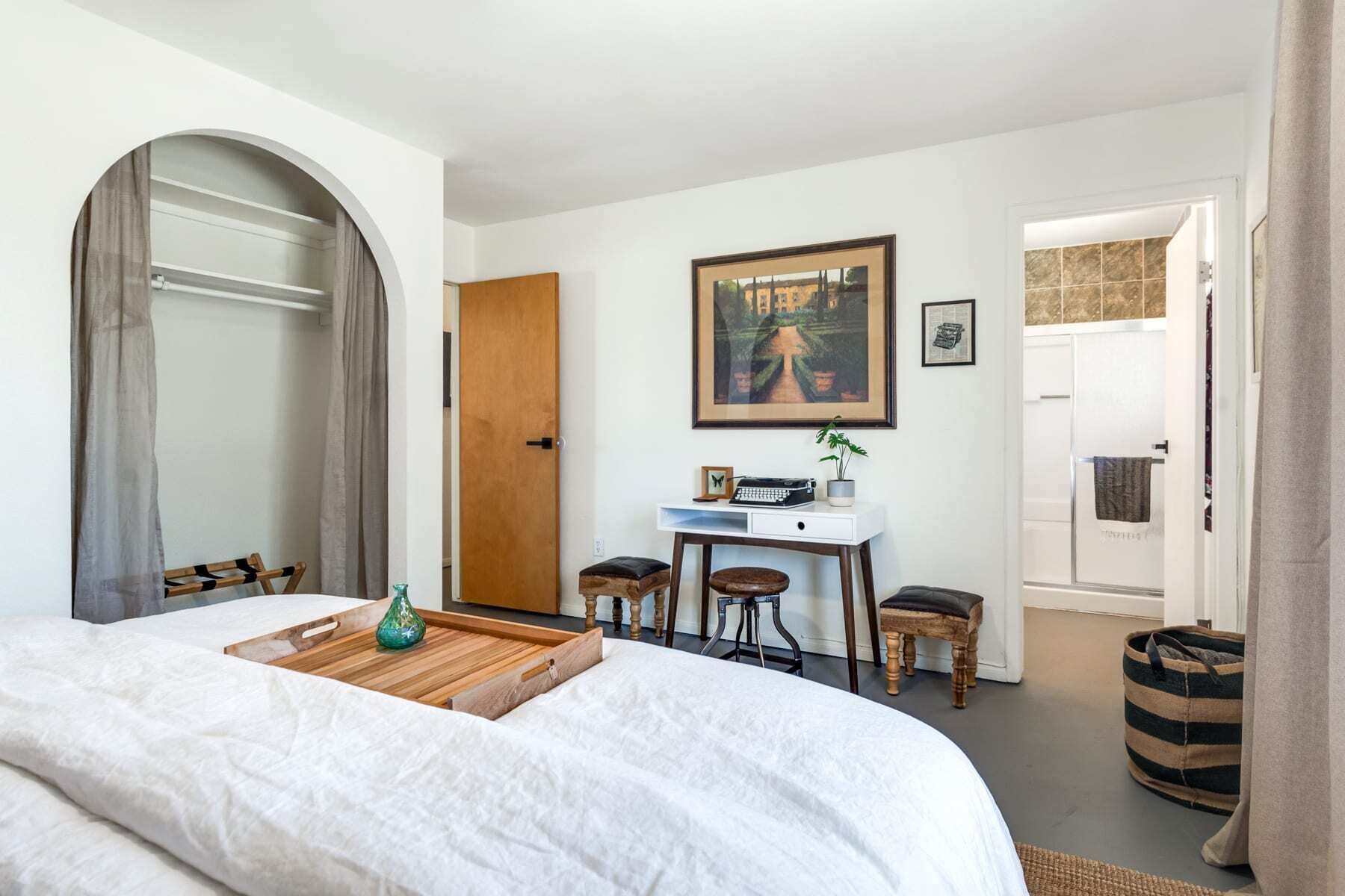 A bedroom with a bed, a desk, two chairs, a wooden side table, a framed picture, and a door leading to a bathroom.