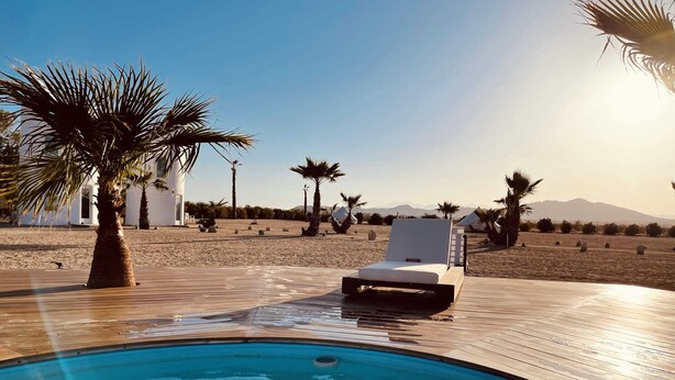 A poolside area with a lounge chair, palm trees, and a clear view of the beach and mountains in the background. - The Castle House: Hideaway w Cowboy Pool (Joshua Tree)