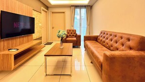 A modern living room with a flat-screen TV, a wooden entertainment unit, a brown leather sofa, a tufted armchair, and a coffee table with a vase of flowers.