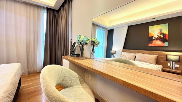 A modern hotel room with a wooden desk, a comfortable armchair, a bed, and a large mirror reflecting the room.