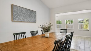 A dining room with a wooden table, six chairs, a vase with greenery, a framed abstract artwork, and large windows with blinds.
