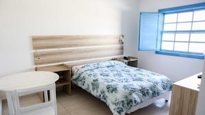 A bedroom with a wooden headboard, a bed with a floral bedspread, a bedside table, a small white table, and a window with blue shutters. - Rafas Pousada Encantos de Minas  (Tres Coracoes)