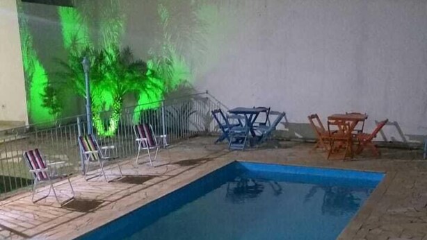 A rectangular swimming pool with a stone border and a metal fence. There are lounge chairs and tables around the pool. - Rafas Pousada Encantos de Minas  (Tres Coracoes)