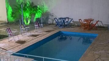 Piscina ao ar livre com deck de pedra, cercada por uma grade de metal, e equipada com espreguiçadeiras e mesas.