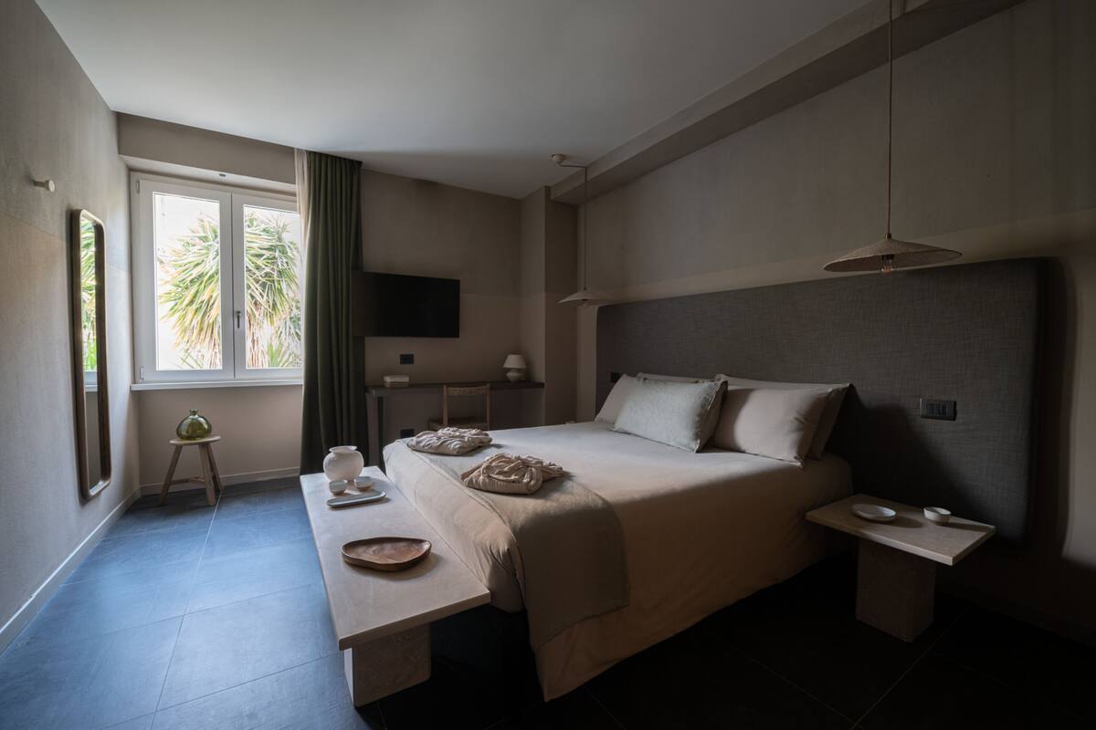 A modern hotel room with a large bed, bedside tables, a small table with a bowl, a TV on the wall, and a window with curtains.