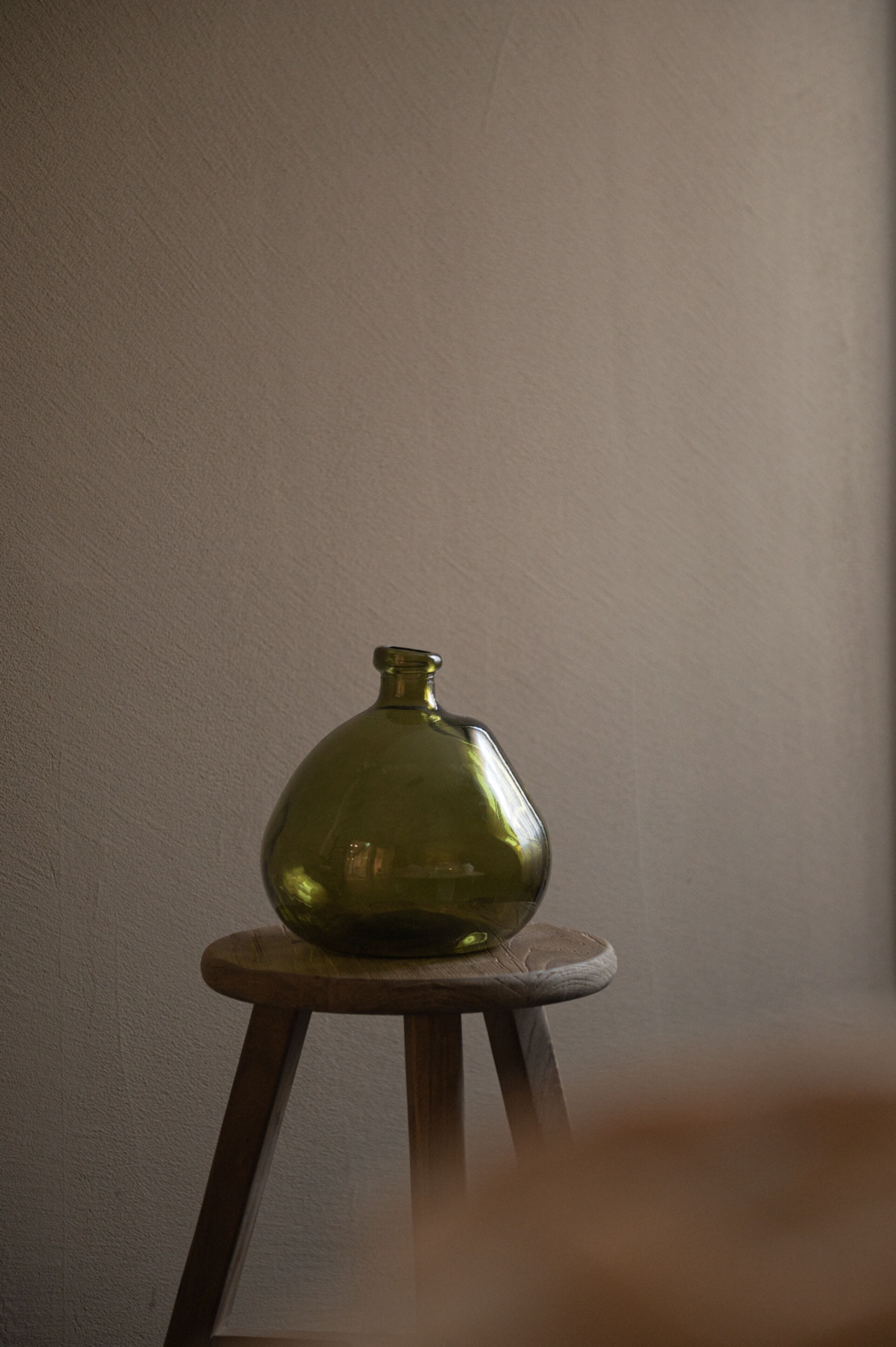 A green vase on a wooden stool against a plain wall.