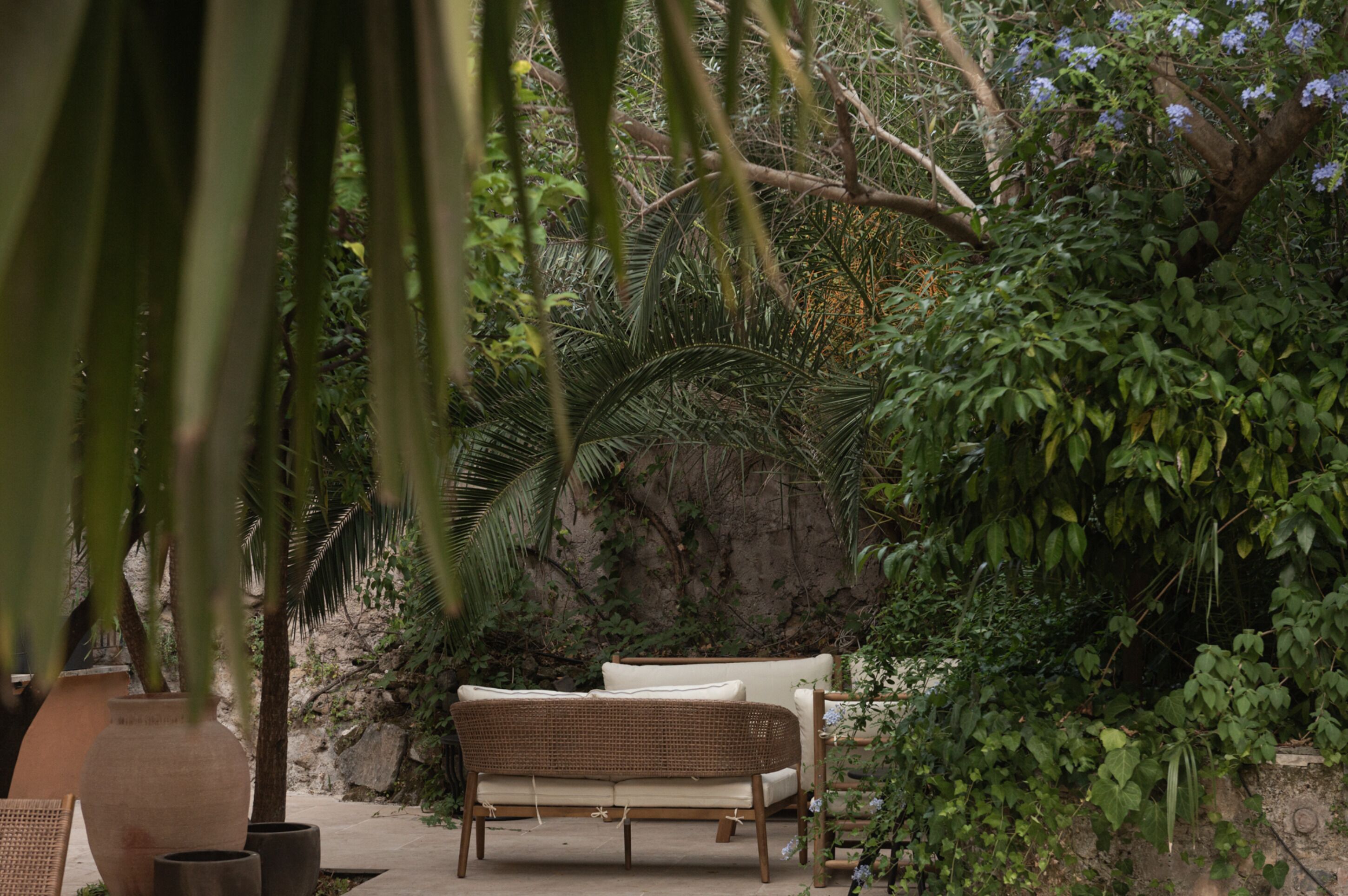 A wicker sofa and a chair in a lush garden setting with a rock wall and various plants.