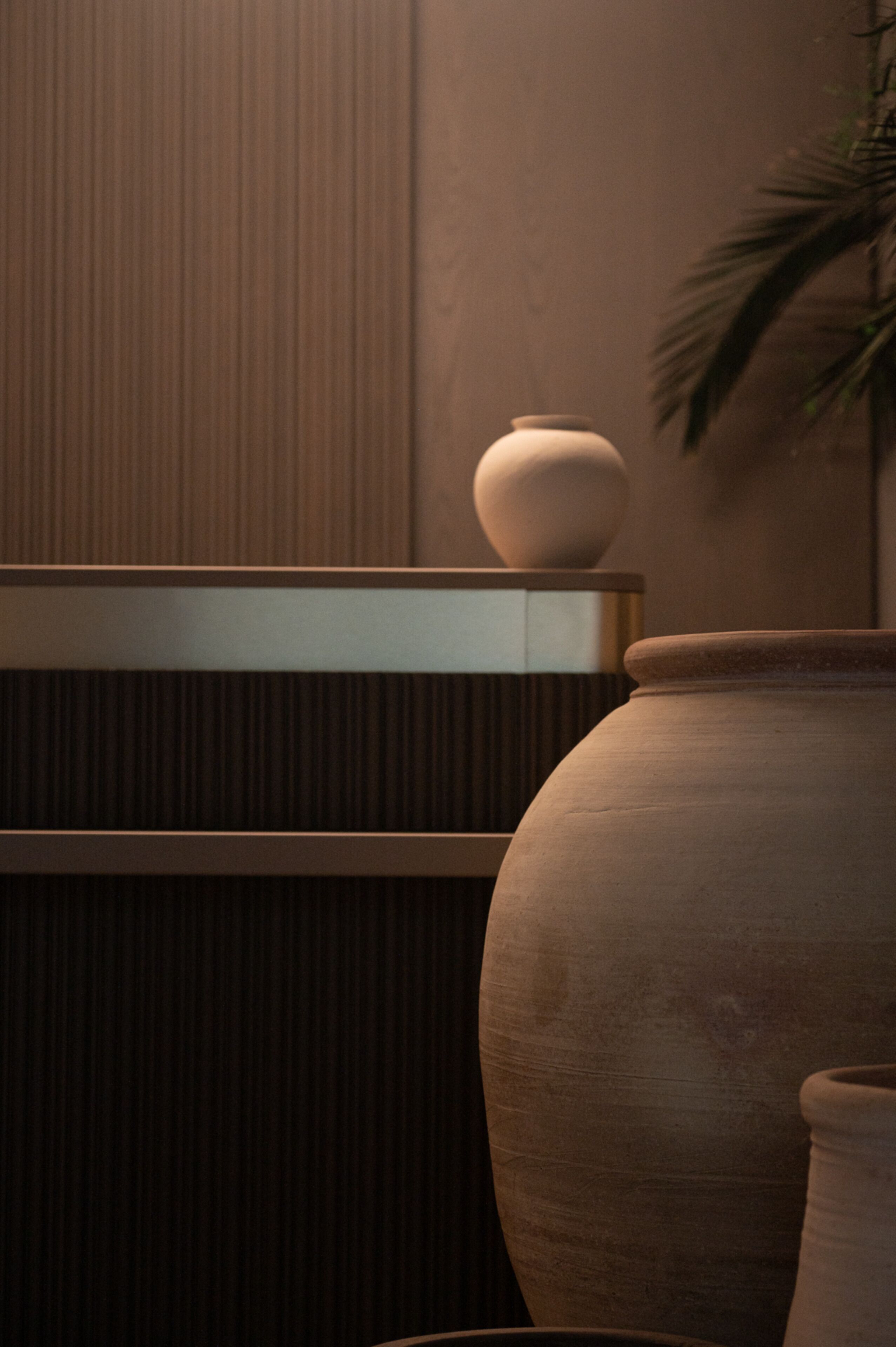 A room with a large, round, beige ceramic vase on a wooden shelf, a smaller vase on the floor, and a potted plant to the right.