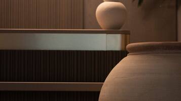 A room with a large, round, beige ceramic vase on a wooden shelf, a smaller vase on the floor, and a potted plant to the right.