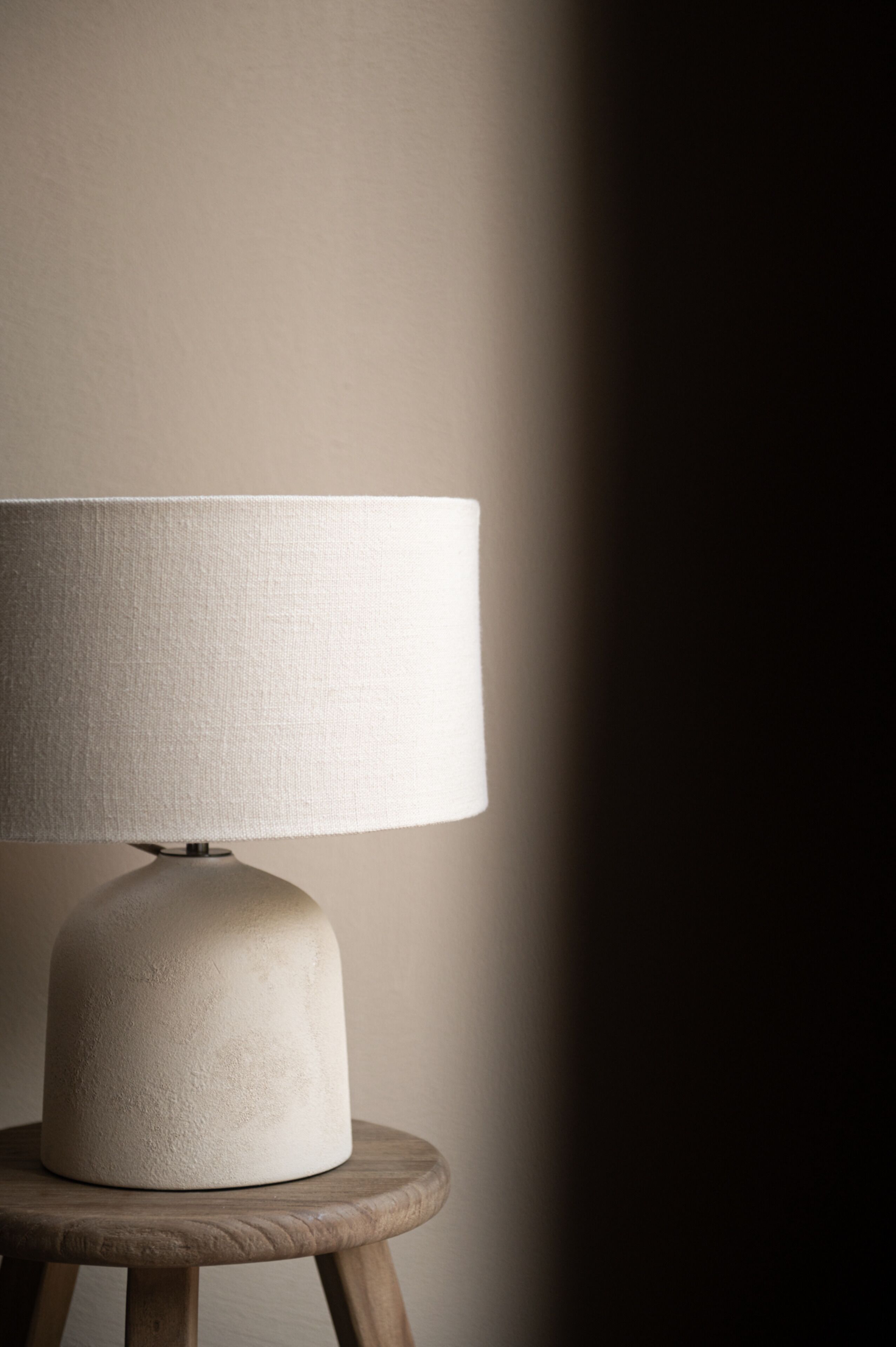 A table lamp with a white shade on a wooden stool against a plain wall.