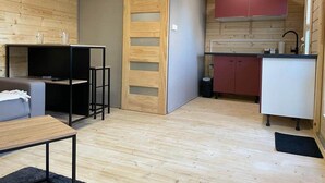 Ein kleines Zimmer mit Sofa, einem Tisch, Fernseher, einem Holzschrank und einer Küchenzeile mit roten Schränken und einem weißen Kühlschrank.