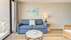 A living room with a blue sofa, a small table with a book, a wicker chair, and a painting of a beach scene. - 1010 Relax, Unwind, Enjoy by Atlantic Towers (Carolina Beach)