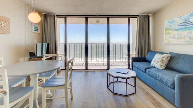 A room with a dining table, chairs, a sofa, a small coffee table, and a large sliding glass door leading to a balcony with a view of the ocean. - 1010 Relax, Unwind, Enjoy by Atlantic Towers (Carolina Beach)