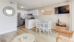 A modern kitchen with white cabinets, a central island, and a dining area with white chairs. - 1010 Relax, Unwind, Enjoy by Atlantic Towers (Carolina Beach)