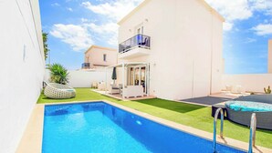 Une piscine dotée d’une balustrade en verre, un espace extérieur aménagé et couvert, et un bâtiment blanc avec un balcon.