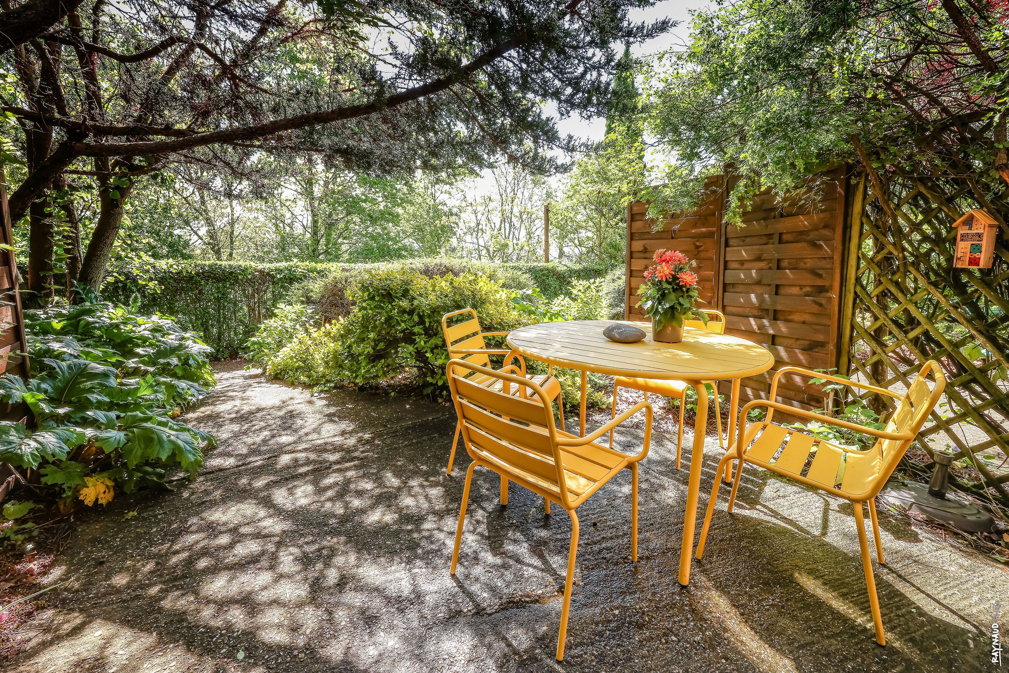 Eine Garten-Terrasse mit einem Holztisch und gelben Stühlen, umgeben von üppiger Bepflanzung und einem Holzzaun.