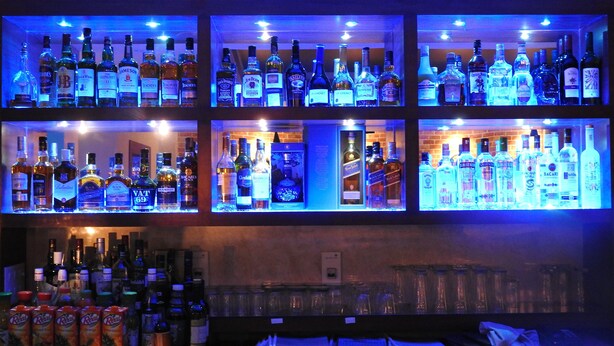 Een bar met meerdere planken vol drankflessen, glazen en een toonbank met verschillende flessen en blikjes.