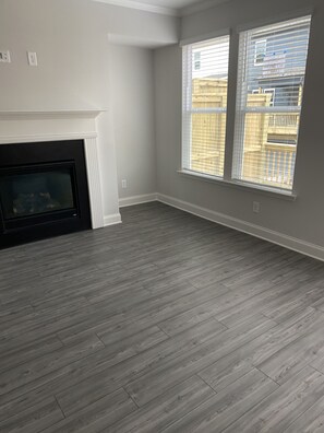A room with a fireplace, large windows, and a grey wooden floor.