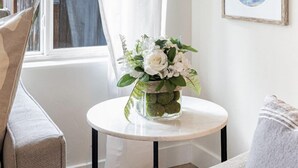 Une pièce dotée d’une grande fenêtre, d’un rideau blanc, d’une table ronde ornée d’un vase de fleurs et de deux tableaux encadrés accrochés au mur.