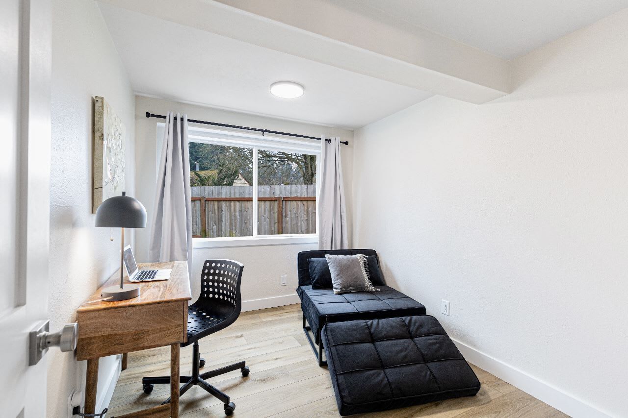A small, bright room with a wooden desk, a black office chair, a bed with a black mattress and pillows, and a window with curtains.