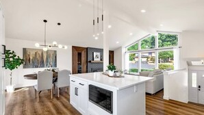Een moderne keuken met een centraal eiland, houten vloeren en grote ramen met uitzicht op groen.