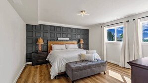 Een slaapkamer met een bed, nachtkastjes, een lamp, een bankje en een raam met gordijnen.