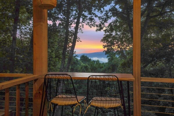 A wooden deck with two metal chairs facing a forested landscape during sunset.