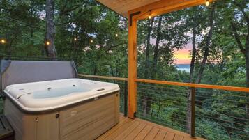 A hot tub on a wooden deck with a view of trees and a sunset.