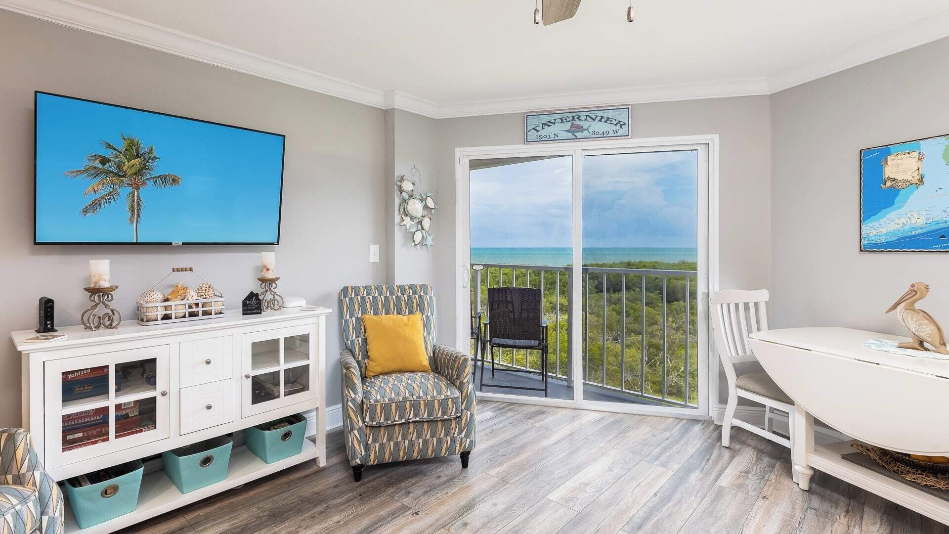 A room with a large TV, a patterned armchair, a dining table, and a view of the ocean through a sliding door.