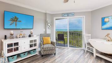 A room with a large TV, a patterned armchair, a dining table, and a view of the ocean through a sliding door.