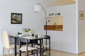 A dining area with a table set for two, a vase with flowers, and a wall-mounted shelf with decorative items.