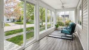 A covered porch with a view of a suburban street, a ceiling fan, and a single teal chair. - New Remodel with Hot Tub 3 blocks from Beach, 1 block to Town, & Game Room! (South Haven)