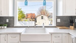 A kitchen with a large window, white cabinets, a sink, and hanging blue pendant lights. - New Remodel with Hot Tub 3 blocks from Beach, 1 block to Town, & Game Room! (South Haven)
