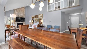 A spacious dining area with a long wooden table, comfortable seating, a stone fireplace, and a balcony with a railing. - Family and Pet Friendly Mountain Home with a view.  No Hurricane Damage (Pisgah Forest)