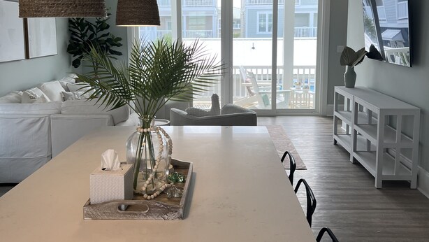 A modern living room with a large white sofa, a dining table with a vase and decorative items, and a view of a balcony with a railing. - NEW home in 2023 w/ elevator! Perfect family getaway in Emerald Isle, sleeps 11! (Emerald Isle)