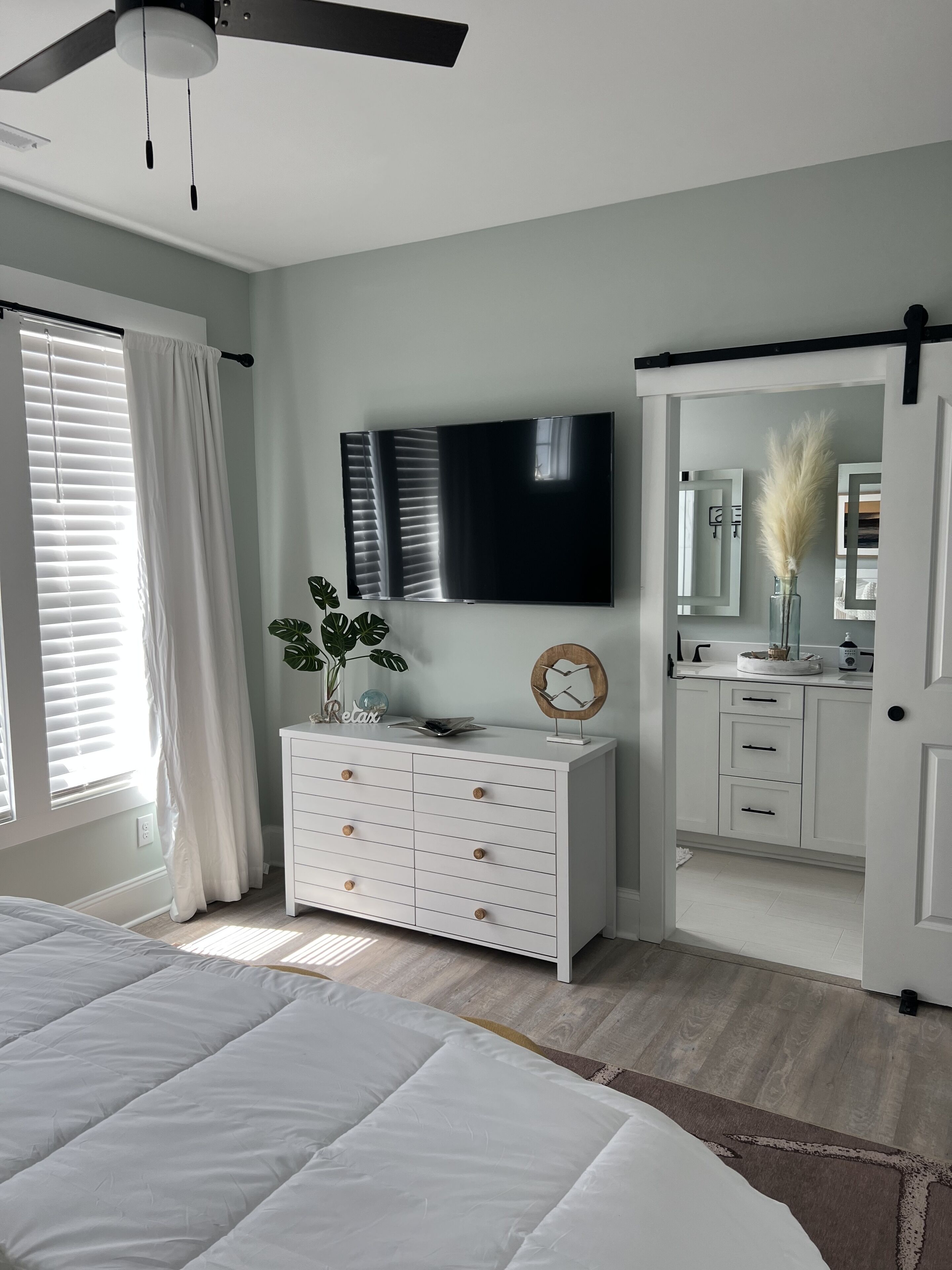 Une chambre à coucher comprenant un lit, une commode, un téléviseur, un ventilateur de plafond, et une salle de bain visible à travers une porte ouverte.