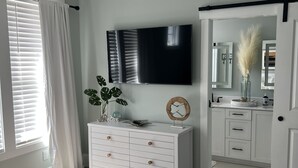 A bedroom with a bed, a dresser, a TV, a ceiling fan, and a bathroom visible through an open door. - NEW home in 2023 w/ elevator! Perfect family getaway in Emerald Isle, sleeps 11! (Emerald Isle)