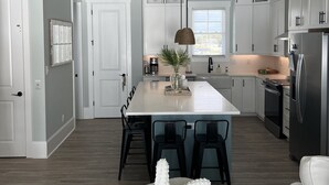 A modern kitchen with white cabinetry, a central island, and a dining area with a starfish decoration. - NEW home in 2023 w/ elevator! Perfect family getaway in Emerald Isle, sleeps 11! (Emerald Isle)
