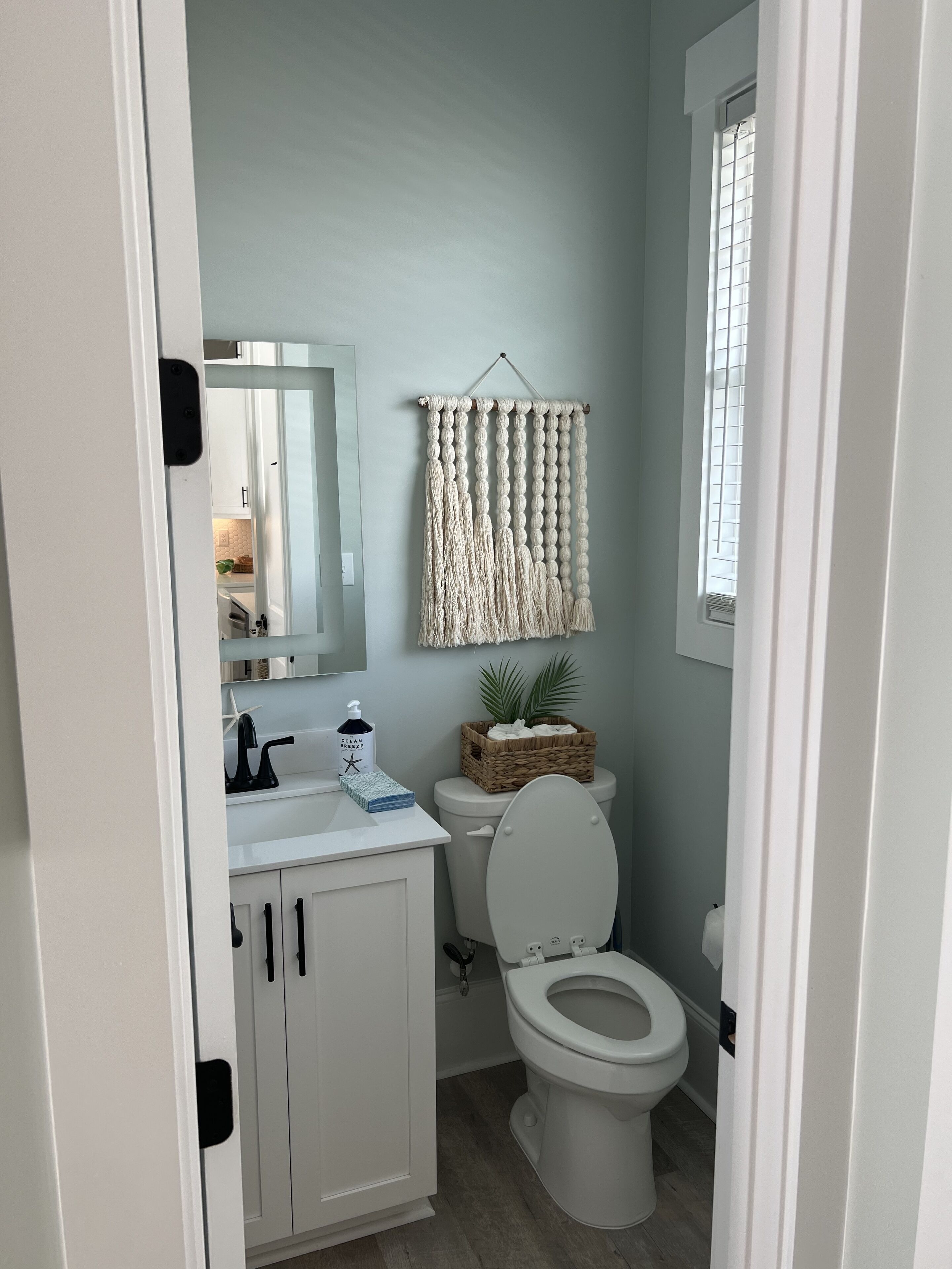 A bathroom with a toilet, sink, and cabinets. There is a macrame wall hanging above the toilet and a basket on the sink.