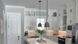 A modern kitchen with white cabinets, a large island with bar stools, pendant lights, and a stainless steel refrigerator. - NEW home in 2023 w/ elevator! Perfect family getaway in Emerald Isle, sleeps 11! (Emerald Isle)