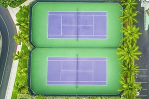 An aerial view of a tennis court complex with two courts, surrounded by a green fence and palm trees.