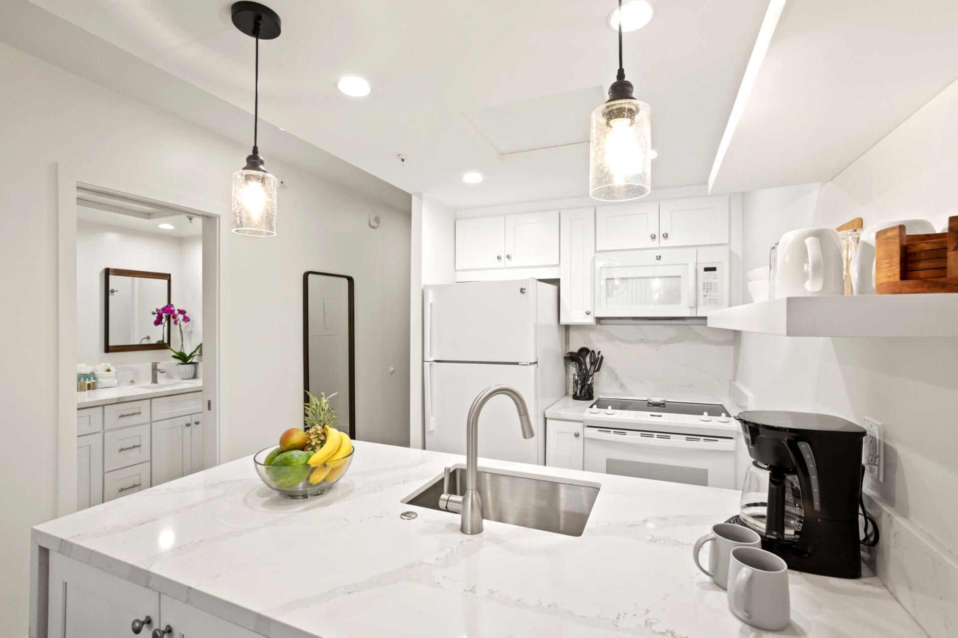 Una cocina moderna con armarios blancos, un lavamanos de acero inoxidable, un frutero y una cafetera.
