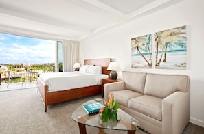 A hotel room with a bed, a sofa, a glass coffee table, and a painting of a beach scene.